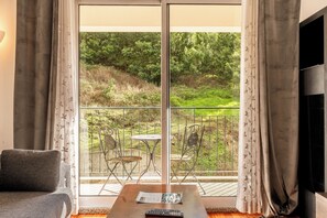 Een woonkamer met een bank, een tafel met een afstandsbediening en een televisie. Er is een balkon met een tafel en stoelen, en uitzicht op groen.