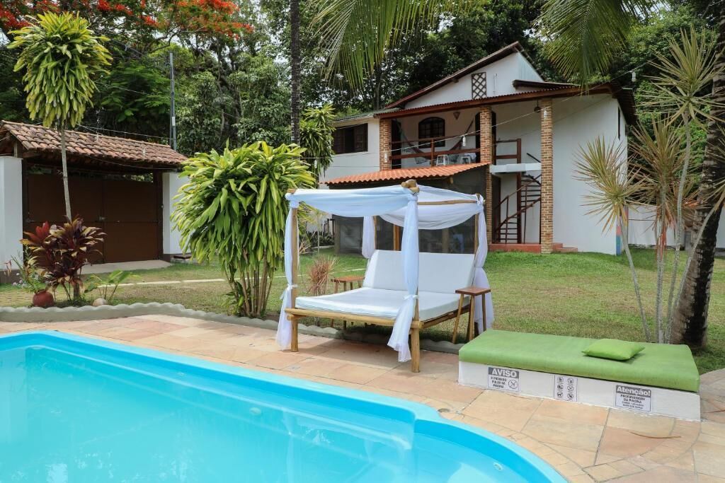 A pool area with a lounge chair and a small table, surrounded by greenery and a house in the background.