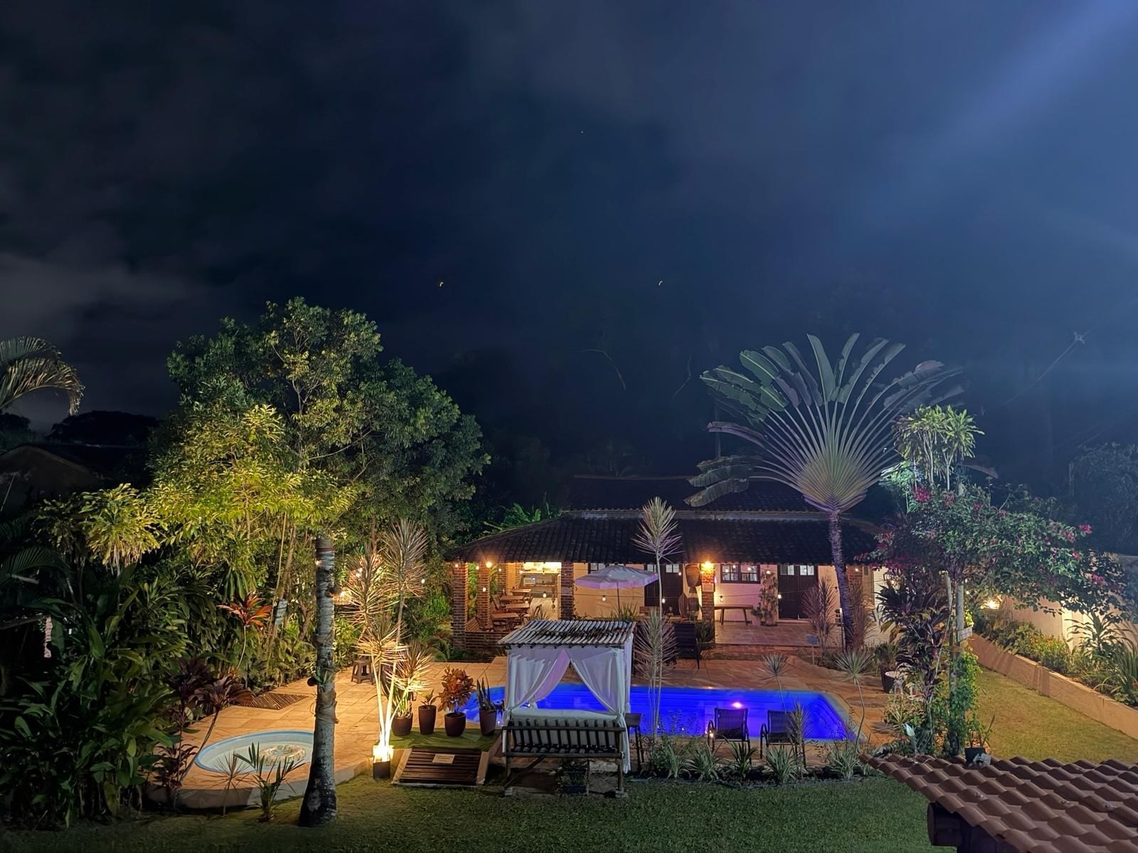 Un espacio exterior bien iluminado con piscina, mobiliario y un edificio de techo con tejas.