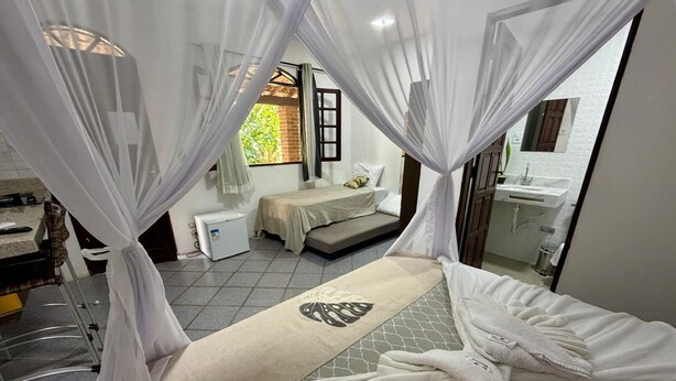 A bedroom with a bed, a small bedside table, a window with curtains, a bathroom with a sink and mirror, and a tiled floor. - Bella vida274 (Porto Seguro)