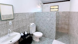 A bathroom with a white sink, toilet, and shower area, marble walls, and a wooden ceiling beam. - Bella vida274 (Porto Seguro)