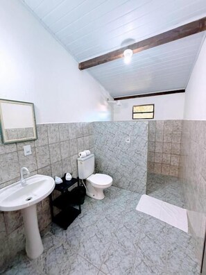 Un baño con lavamanos, inodoro y zona de ducha blancos, paredes de mármol y una viga de techo de madera.