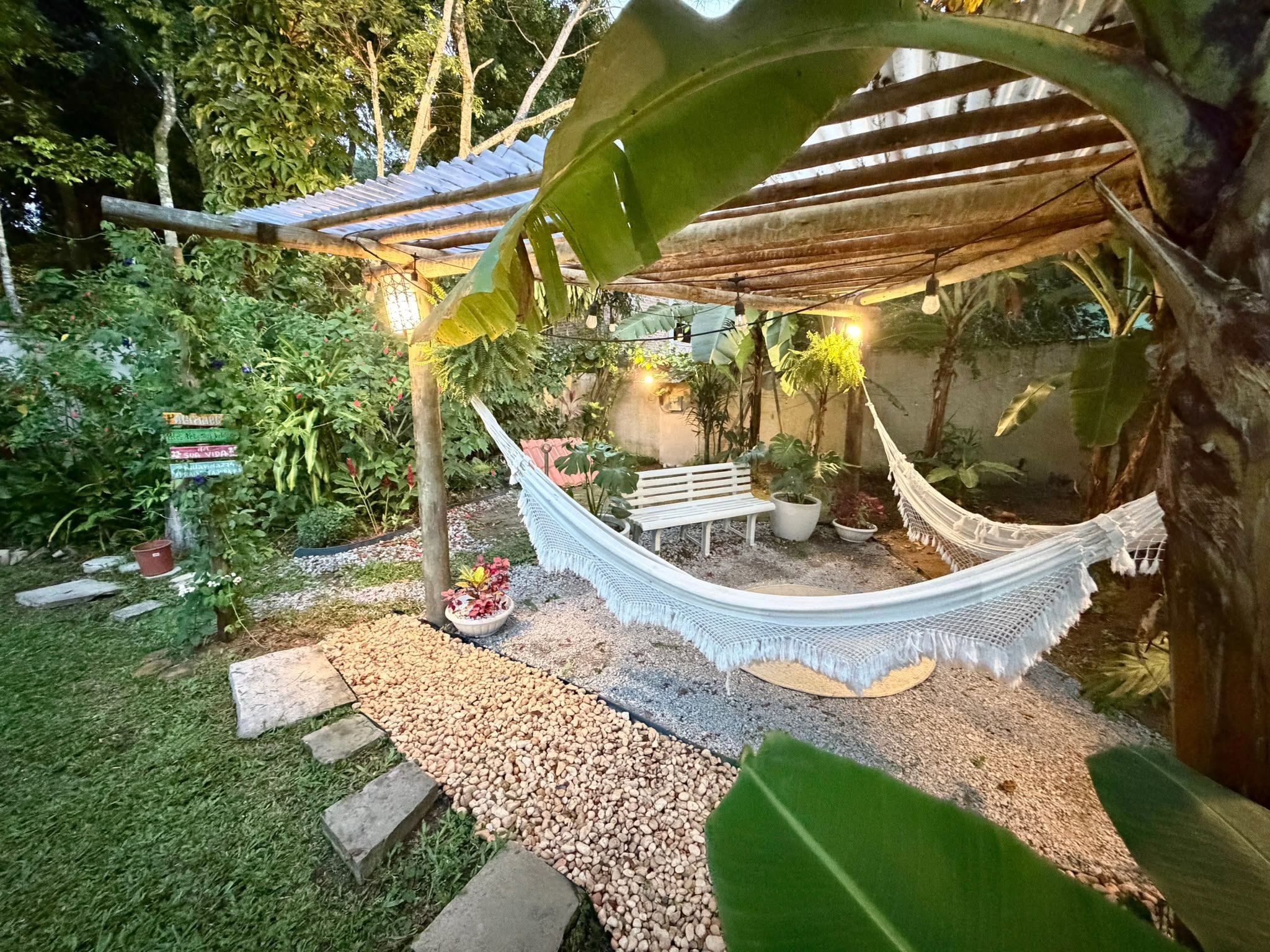 A hammock under a wooden pergola in a garden with a gravel path and potted plants.