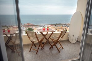 Un balcón con mesa y sillas, una tabla de surf y vistas al mar y edificios.