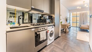 A modern kitchen with a built-in oven, microwave, and sink. There is a dining area with a table and chairs, and a large window providing natural light. - Tranquil Studio with City View, O10 Building (Dubai)