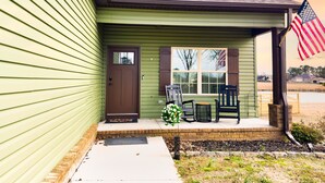 Uma casa com fachada verde, porta da frente marrom e varanda com duas cadeiras e uma pequena mesa.