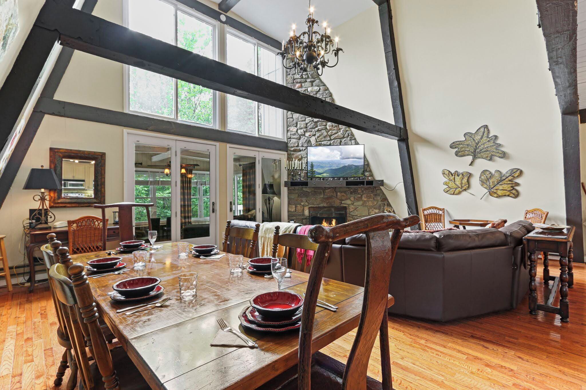 A dining area with a wooden table set for four, surrounded by wooden chairs. A stone fireplace with a chimney is visible in the background, and there is a large window providing natural light.