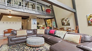 Een ruime zithoek met een grote leren hoekbank, een rond pouf en een vloerkleed met een patroon. Achtergrond is een keuken zichtbaar, en er bevindt zich een balkon met een hekwerk boven de zithoek.