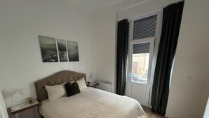 A bedroom with a bed, bedside tables, a radiator, a window with curtains, and wall art. - Ground Serenity (Saint-Gilles)
