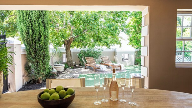 A dining area with a view of a backyard patio, a wooden table, a bowl of fruit, and wine glasses. - Winelands Mountain View Oasis - 3 Bedroom House, Gym, Pool, Cinema, Lounge, BBQ (Franschhoek)