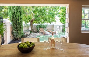 A dining area with a view of a backyard patio, a wooden table, a bowl of fruit, and wine glasses. - Winelands Mountain View Oasis - 3 Bedroom House, Gym, Pool, Cinema, Lounge, BBQ (Franschhoek)