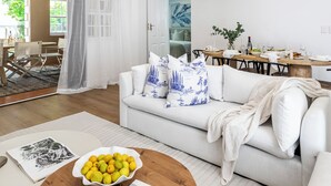 A modern living room with a white sofa, a round wooden coffee table with a bowl of lemons and a magazine, and a dining area in the background. - Winelands Mountain View Oasis - 3 Bedroom House, Gym, Pool, Cinema, Lounge, BBQ (Franschhoek)