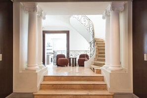 A grand staircase with white columns and a wrought iron railing, leading to a landing with a red upholstered chair and a small table. - Stunning private hotel with rooftop terrace - Neuilly (Neuilly-sur-Seine)
