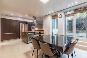 Eine moderne Küche mit Essbereich, ausgestattet mit einem Glastisch, Stühlen, Edelstahlgeräten und Blick durch große Fenster.