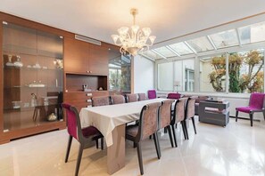 Un espace de repas moderne avec une cuisine ouverte derrière une paroi de verre, un lustre et une vue sur les arbres à travers de grandes fenêtres.