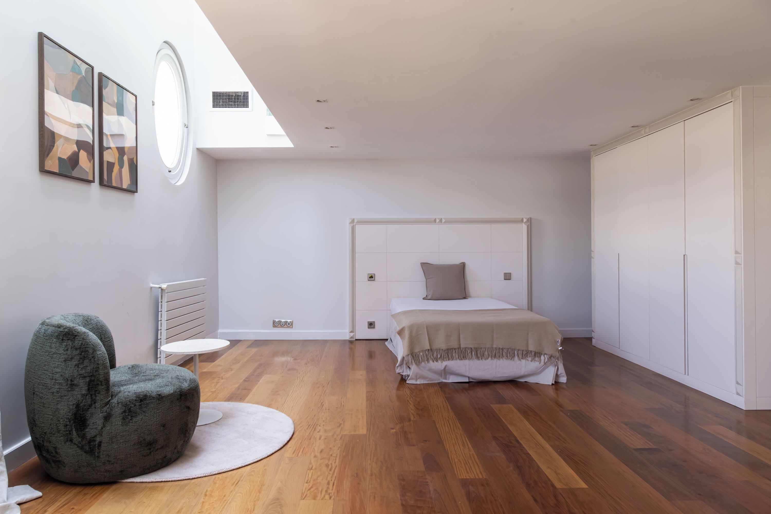 A modern bedroom with a bed, a round chair, a radiator, and artwork on the wall.