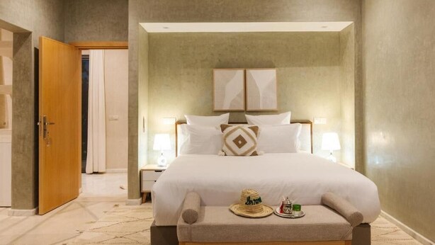 A hotel room with a large bed, bedside tables with lamps, a wooden door, and a small table with a hat and a bottle. - Domaine Almaz 2 Villas and 3 Pools (Aghouatim)