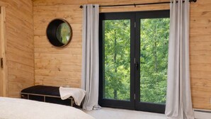 A bedroom with a bed, a small table, a window with curtains, and a view of trees.
