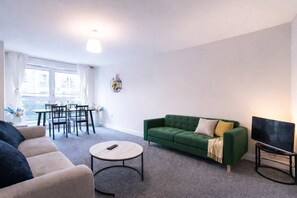 A living room with a green sofa, a beige sofa, a round coffee table, a TV on a stand, and a dining area with a table and chairs. - Spacious 2BR Flat - Bounds Green Private Parking (London)
