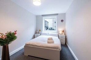 A modern hotel room with a large bed, bedside tables, a window with a view of buildings, and a vase with flowers. - Spacious 2BR Flat - Bounds Green Private Parking (London)