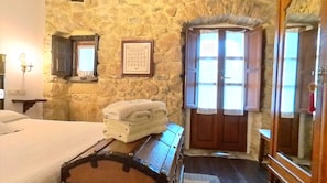 A stone-walled room with a bed, a wooden chest, and a framed picture on the wall.