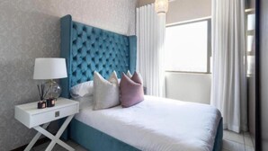 A bedroom with a tufted headboard, a white nightstand with a lamp, and a view of the outdoors through a window with curtains. - The Steynview Apartment (Sandton)