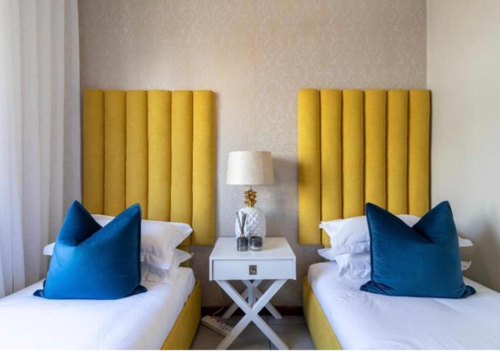 A hotel room with two beds, each with a blue pillow, a white nightstand with a lamp, and a yellow headboard.