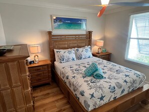 Ein Schlafzimmer mit einem hölzernen Bett, einem Nachttisch mit Lampe, einer Kommode und einem Bild einer Strandlandschaft an der Wand.