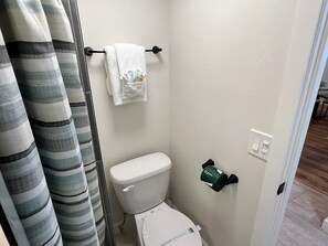 Et lille badeværelse med toilet, et håndklædestativ med et hvidt håndklæde, toiletpapirholder og en badeforhæng.