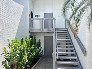 A narrow outdoor corridor with a white brick wall, a metal staircase, and a small garden area with green plants. - 1017 - Premium - Beach Front (Siesta Key)