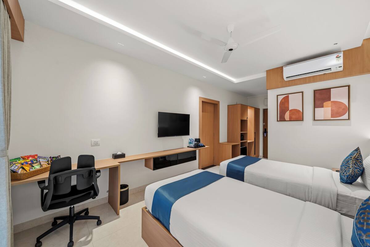 A hotel room with two beds, a desk, a chair, a TV, and an air conditioning unit.