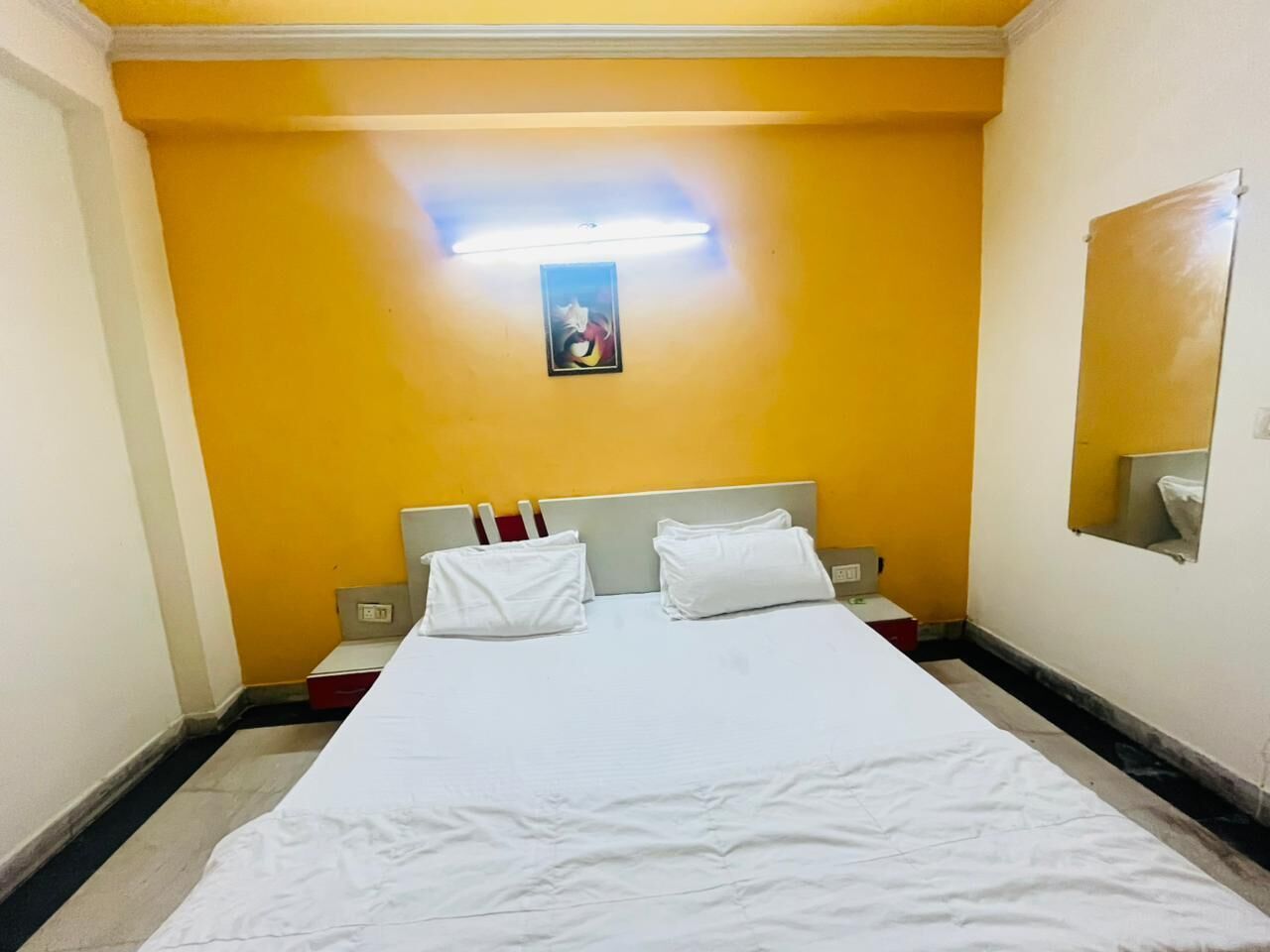 A hotel room with a large bed, white bedding, a single bedside table, a nightstand, a wall-mounted light, and a small framed picture.