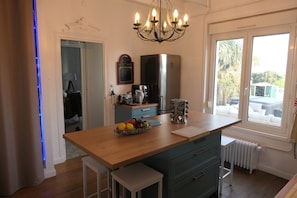 Une cuisine avec un îlot central en bois, un réfrigérateur, un lustre et une fenêtre donnant sur l’extérieur.