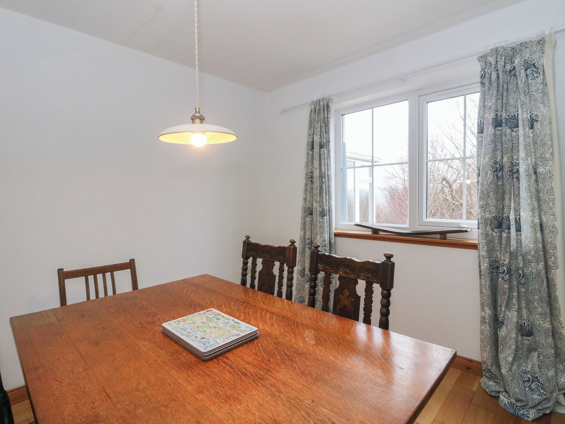 A dining room with a wooden table, chairs, a hanging light fixture, a window with curtains, and a view of trees outside.