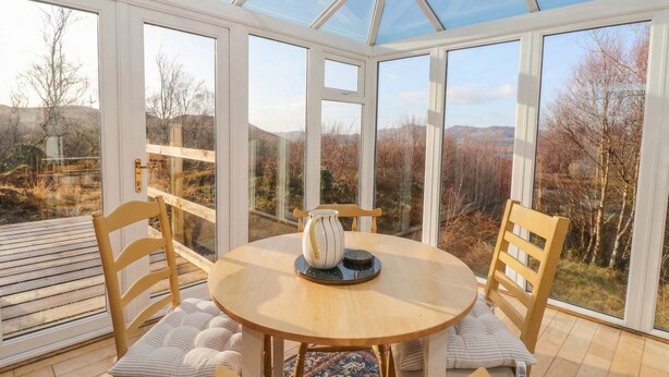 A conservatory with a round wooden table and chairs, large windows, and a view of the countryside. - Doire An Daimh (Acharacle)