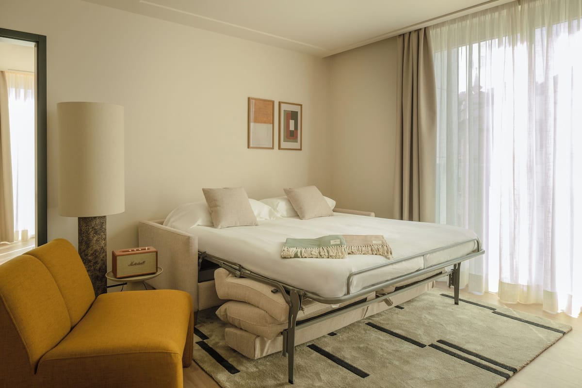 Una habitación de hotel con una cama, una silla, una lámpara y una mesita con un radio.