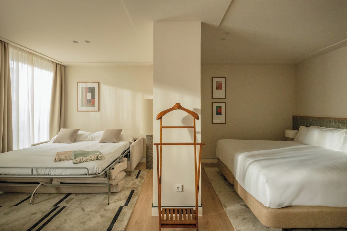 Habitación de hotel con dos camas, un perchero de madera y un ventanal grande con cortinas.