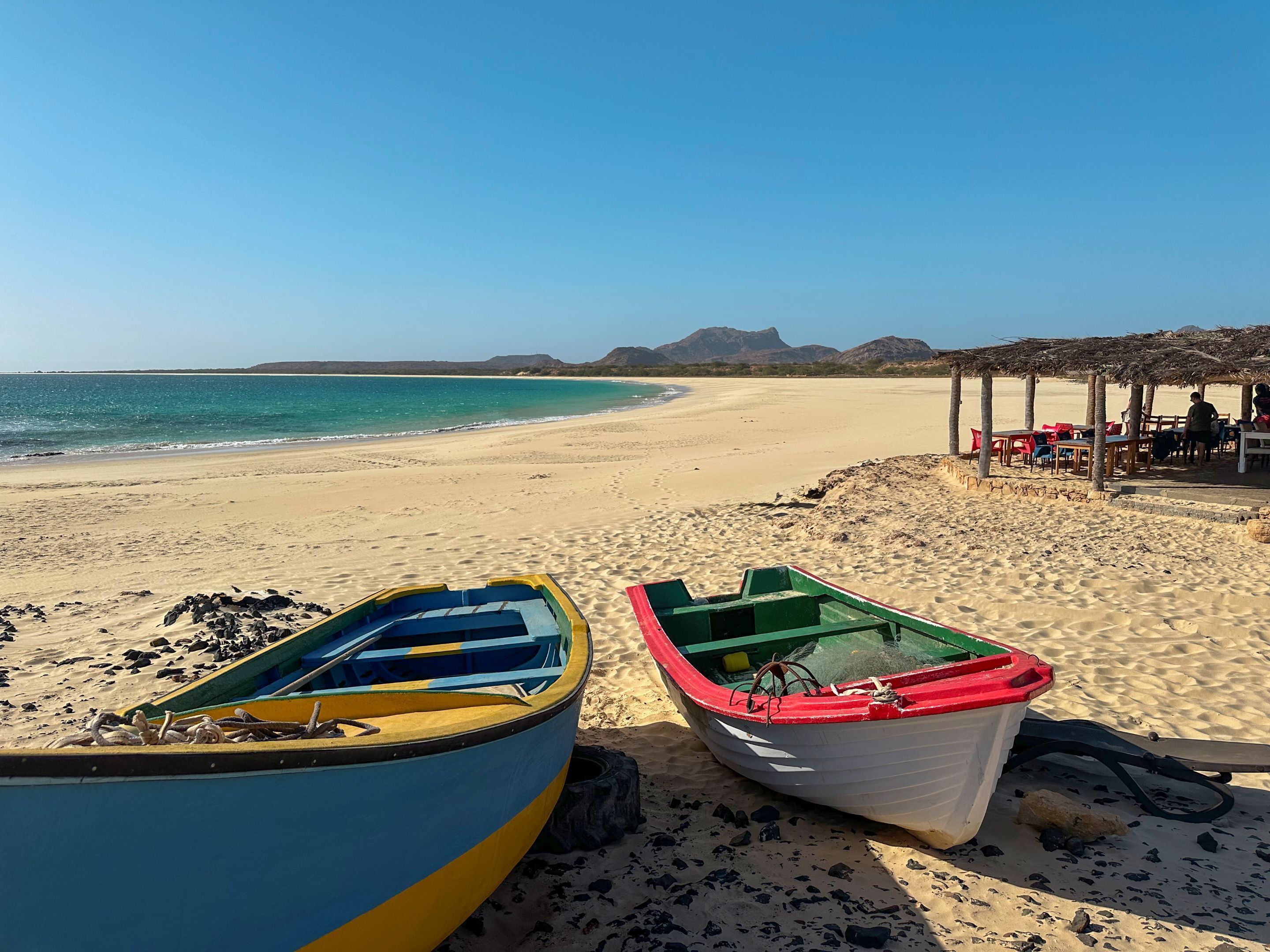 Dos barcos en una playa arenosa con cielo azul despejado y montañas a lo lejos.