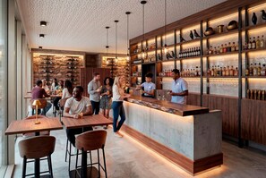 Uma área de bar com um balcão comprido, prateleiras com garrafas e pessoas interagindo.