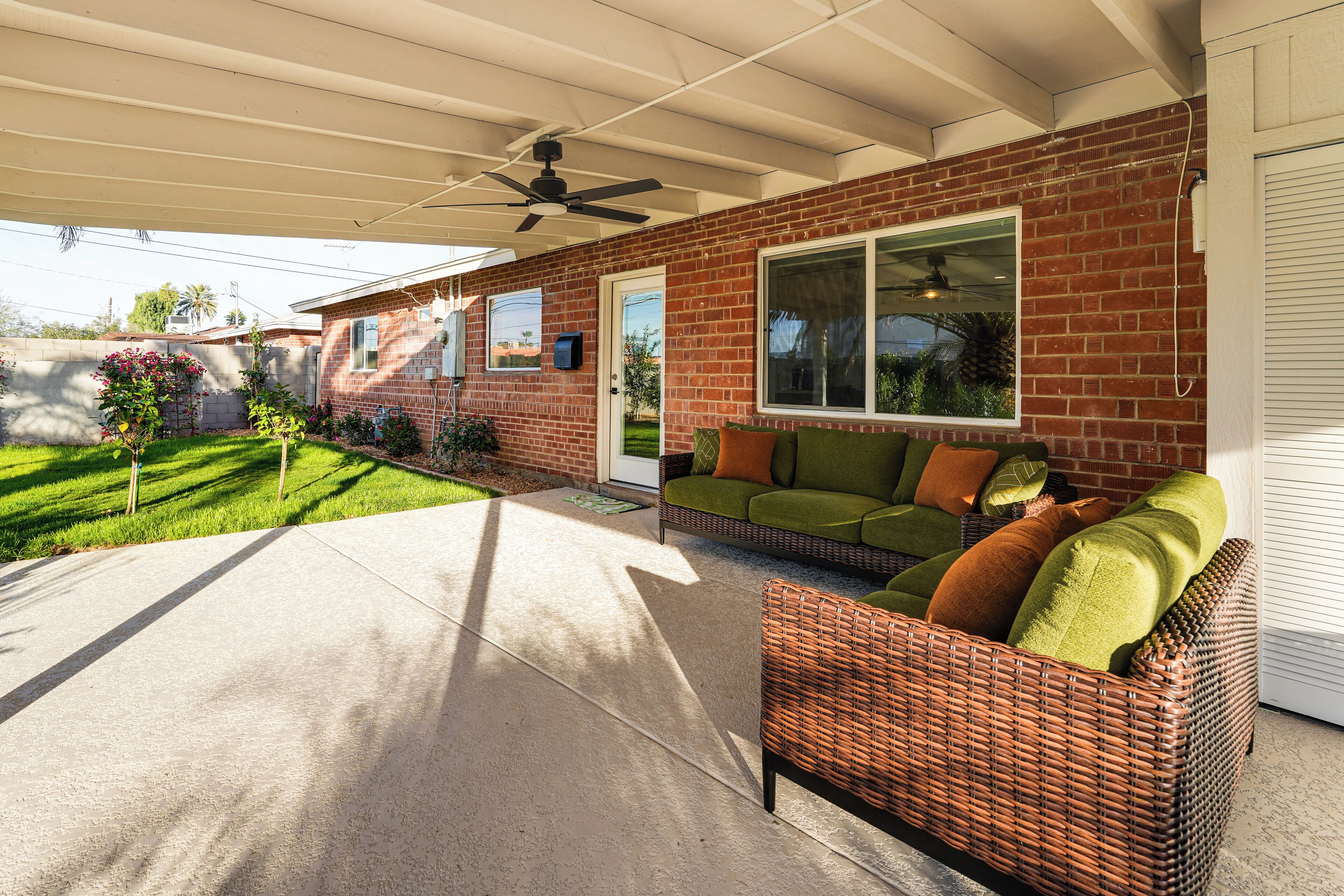 A covered patio area with a wicker sofa and a green cushion, a brick house with a large window, and a small garden with a tree and flowers.