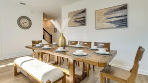 Comedor con una mesa de madera dispuesta para seis personas, rodeada de sillas, un banco y un florero con plumas blancas.