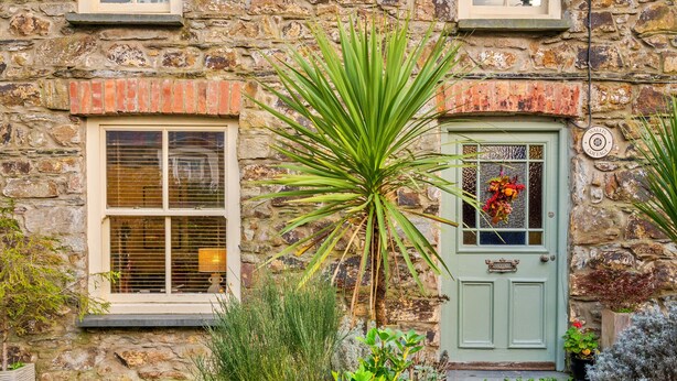 Ein Steinhaus mit einer grünen Tür, einem Fenster mit Rollläden und einem Blumentopf im Vordergrund.
