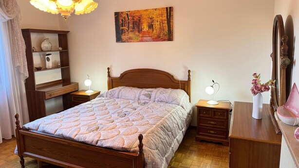 A bedroom with a wooden bed, bedside tables with lamps, a dresser, a mirror, a shelf with decorative items, and a painting on the wall. - La Coccinella Apartment with Better Sleep Technology (Gonars)