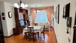 A dining area with a table and chairs, a sofa, a television, wooden cabinets, and a clock on the wall. - La Coccinella Apartment with Better Sleep Technology (Gonars)