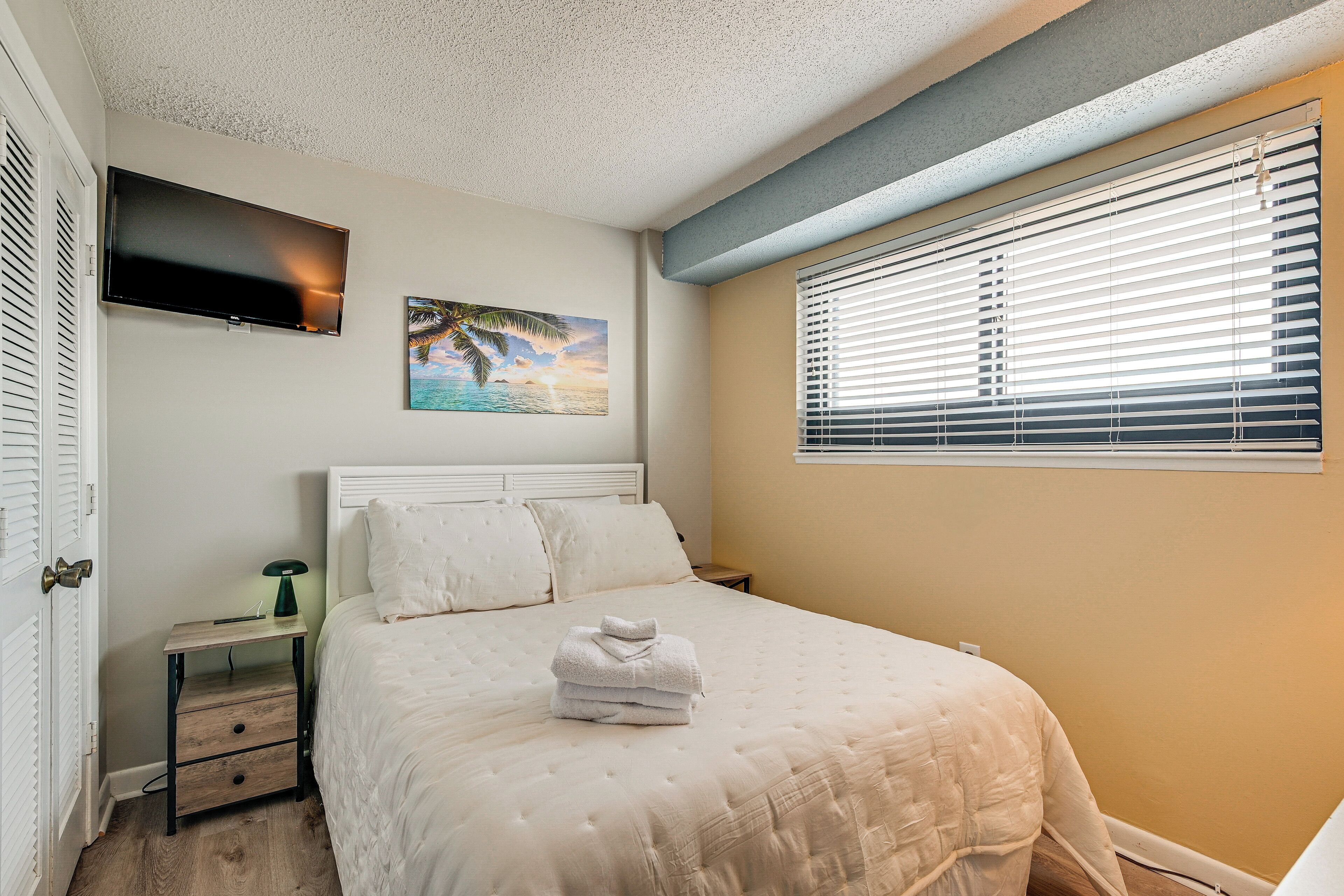 A bedroom with a bed, a nightstand, a TV mounted on the wall, and a window with blinds.