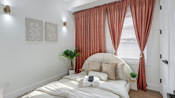 A bedroom with a bed, two wall-mounted light fixtures, a window with curtains, and a potted plant. - 6 Mi to Manhattan: Luxe Apt w/ City Views! (Jersey City)