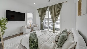 A modern bedroom with a large bed, a flat-screen TV, a seating area with a chair and a small table, and a view of the city through large windows. - 6 Mi to Manhattan: Luxe Apt w/ City Views! (Jersey City)