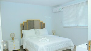Ein Schlafzimmer mit Bett, Nachttischen, Fenster mit Jalousien und einer Klimaanlage.