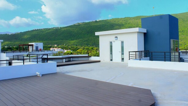 Eine Dachterrasse mit einem modernen Gebäude, einer kleinen Struktur und Blick auf einen grünen Hang.