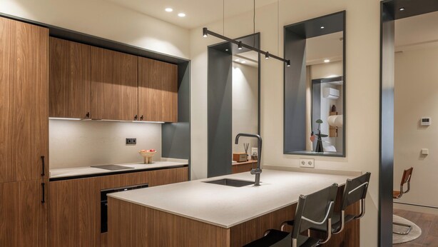 A modern kitchen with wooden cabinets, a central island with chairs, and a large mirror. - POSTCODE Chueca (Madrid)